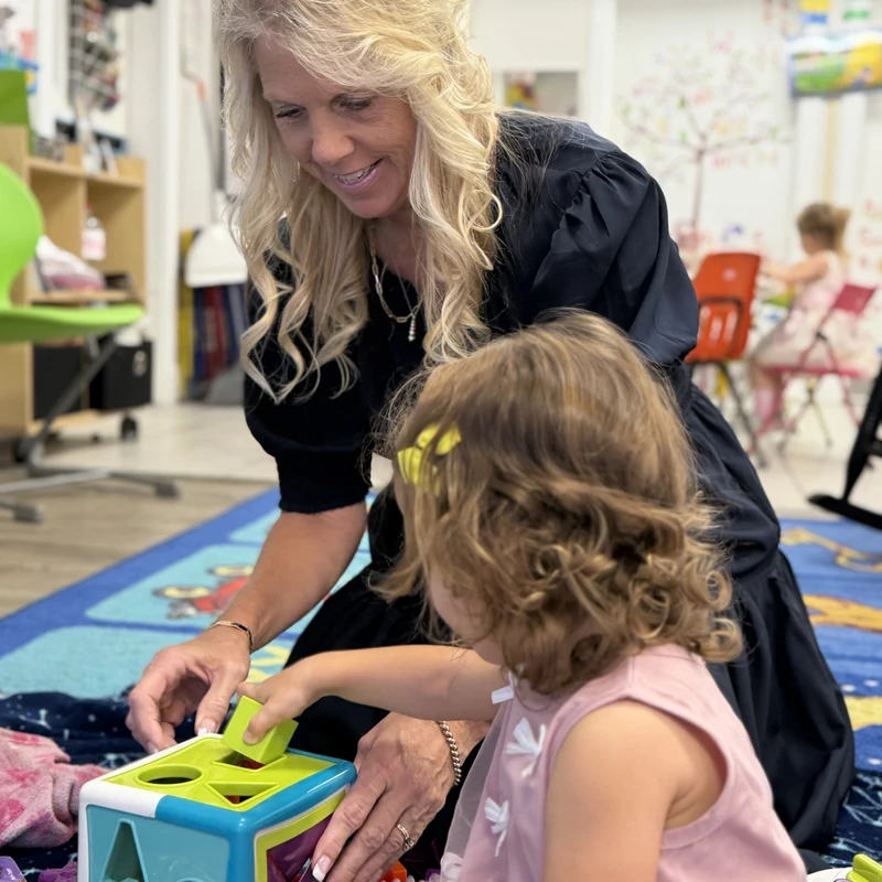 A smiling woman with blonde hair helps a young girl in a pink dress play with a colorful shape sorter toy on a classroom floor.