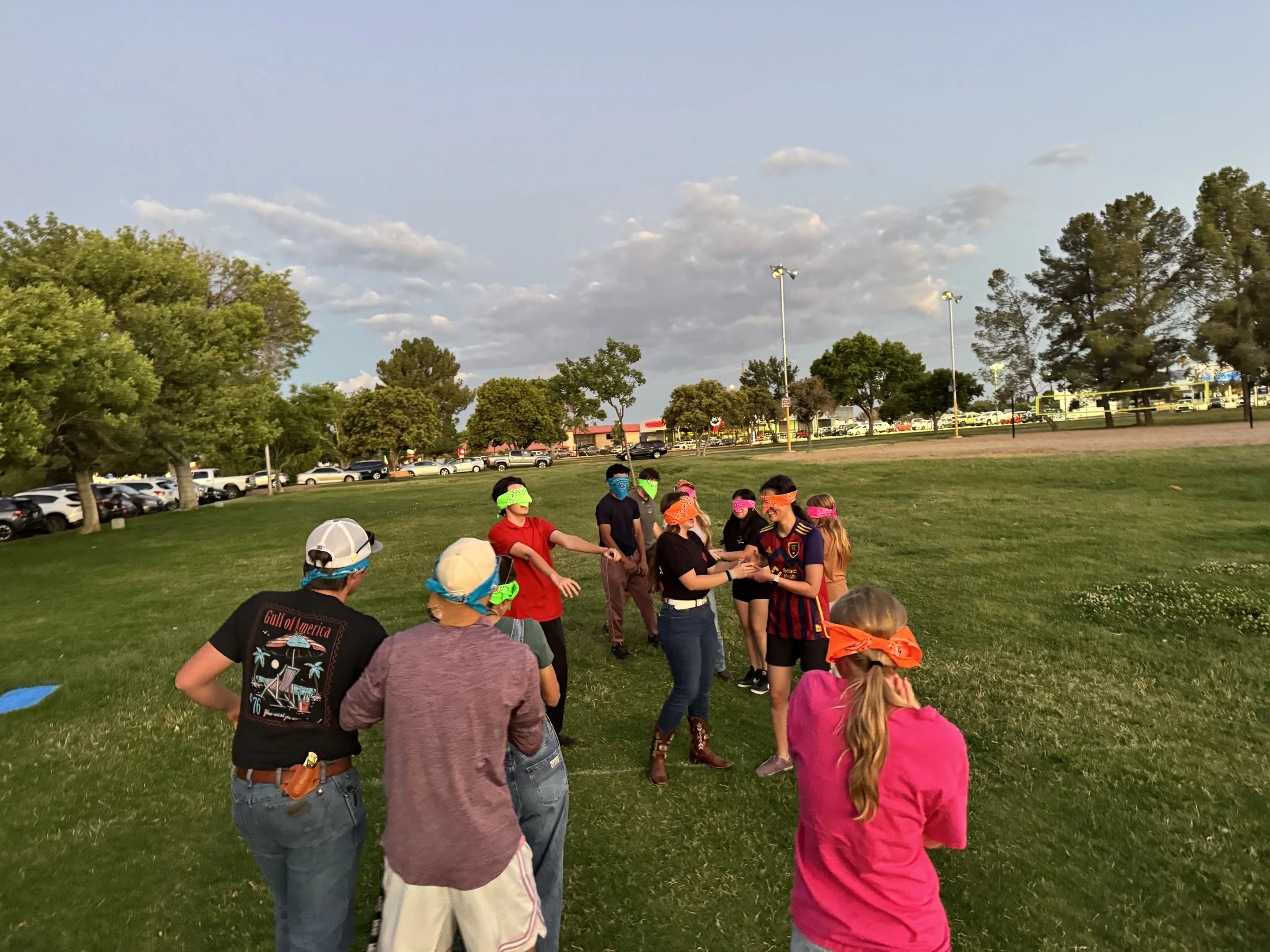 A group of young people wearing colorful bandanas as blindfolds play a game on a grassy field at dusk, with trees and a parking lot in the background.
