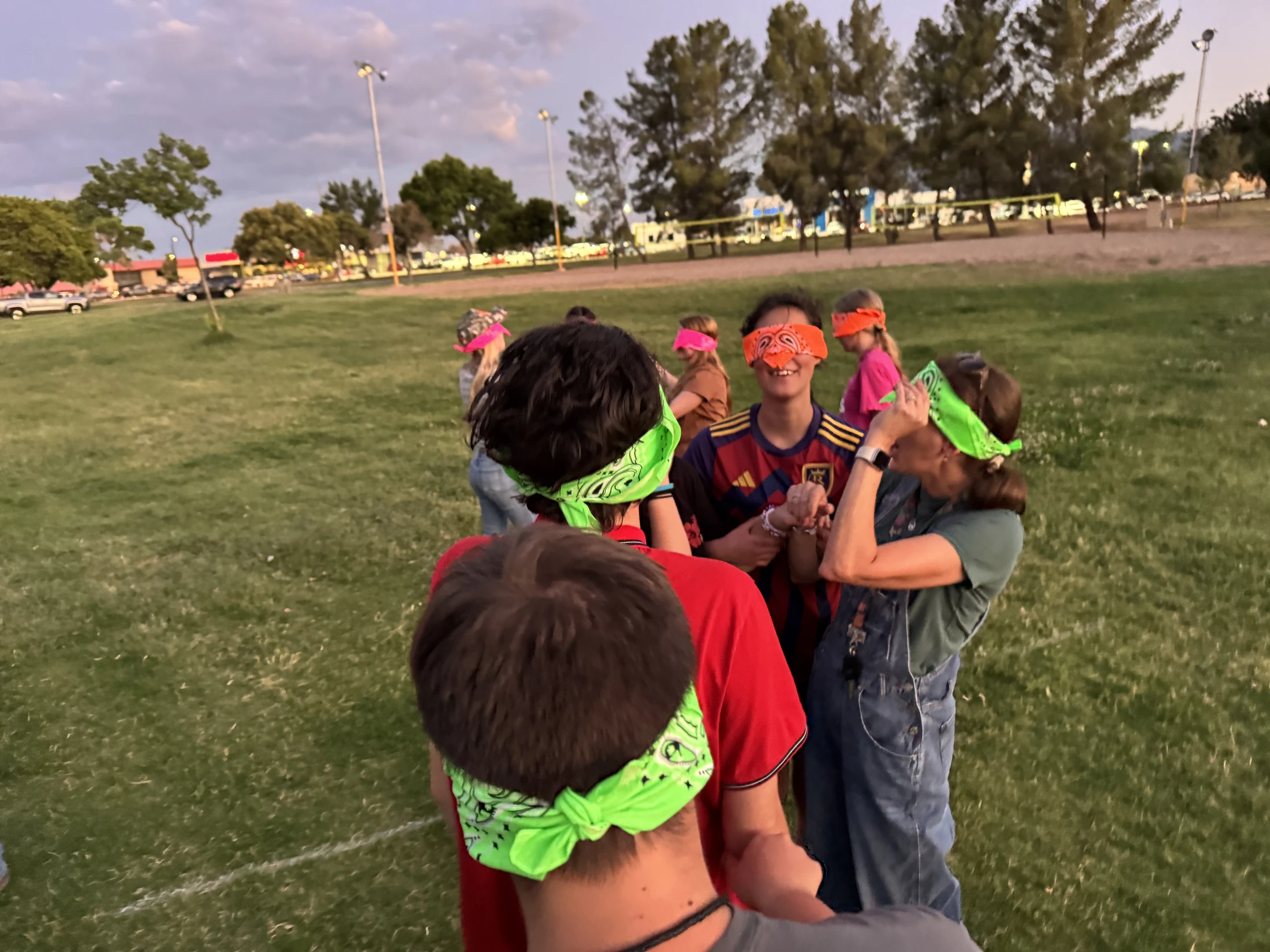 A group of young people stand in a line on a grassy field, wearing colorful bandanas as blindfolds and smiling while playing a game at dusk.