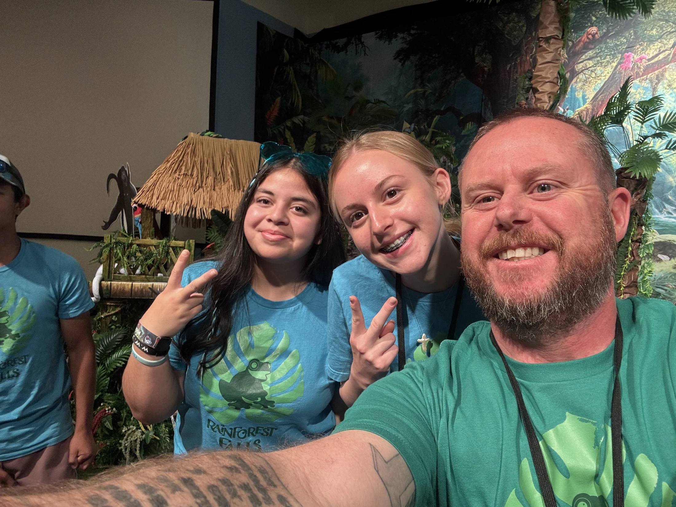 A man with a beard takes a selfie with two smiling young women. They all wear matching 'Rainforest Falls' shirts while flashing peace signs.