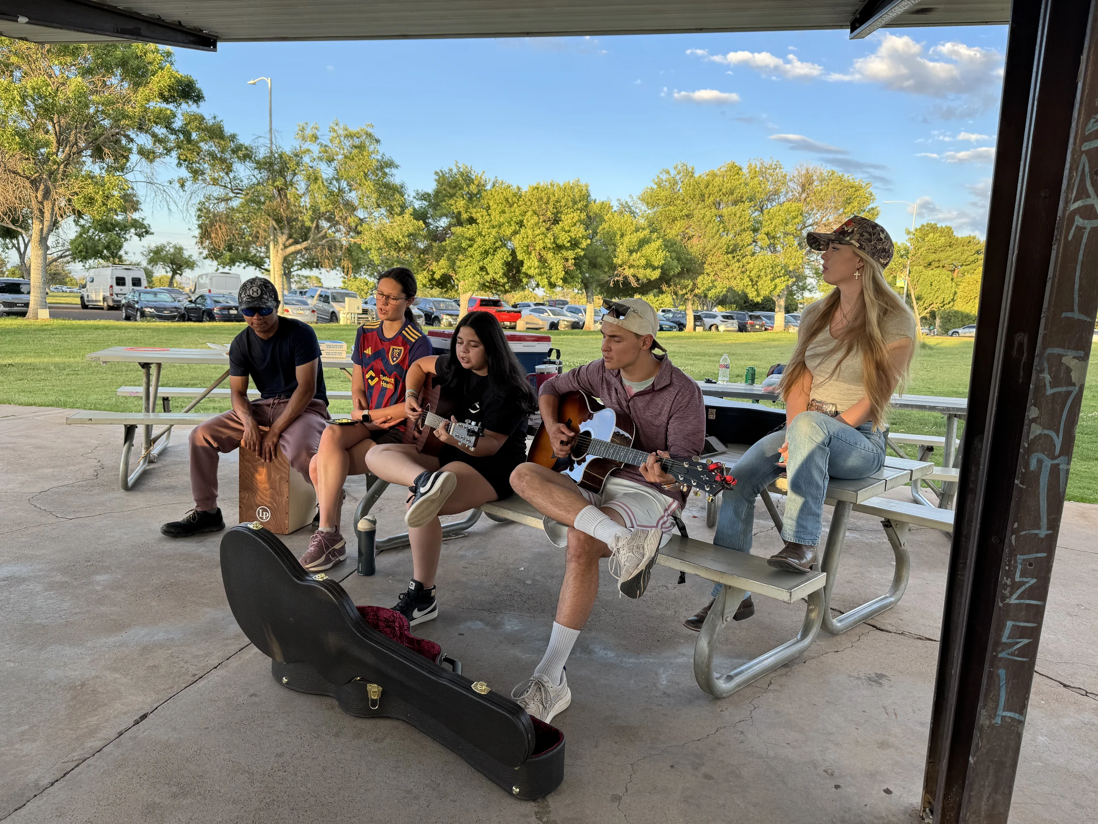 Five young adults sit at a picnic table under a pavilion in a park. Two play acoustic guitars, one plays a cajón drum, and two others listen.