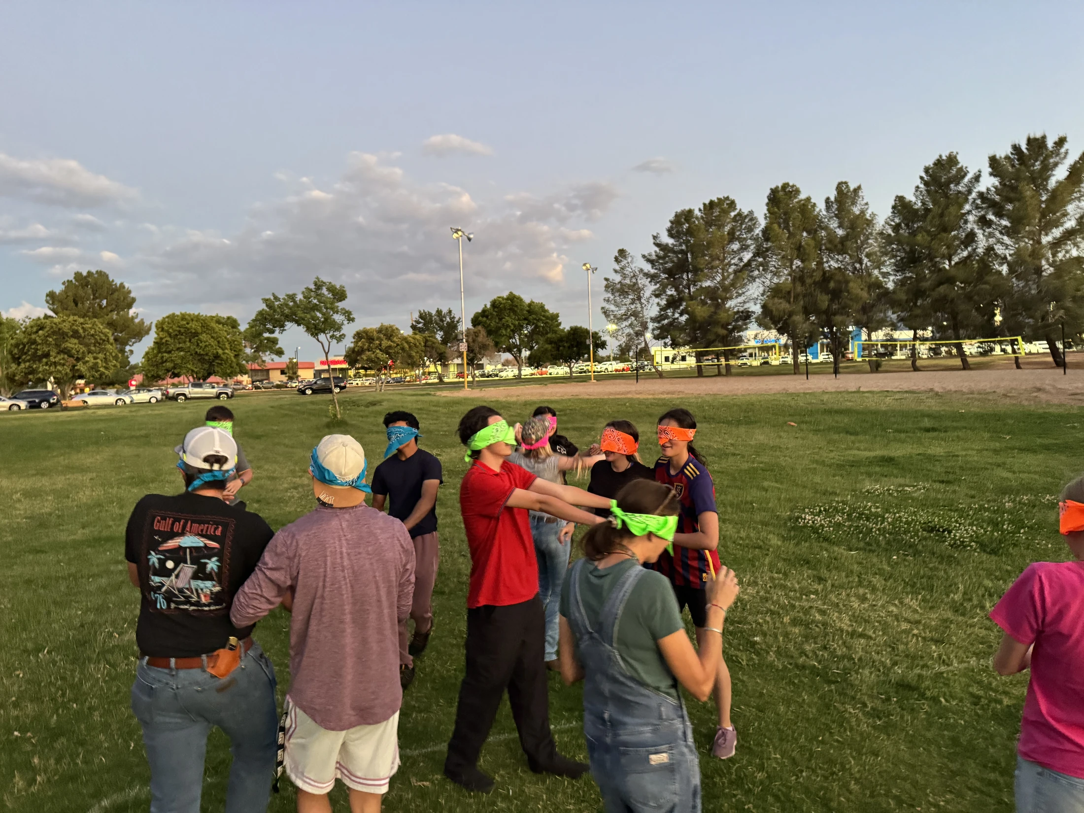 A group of young people stand in a circle on a grassy field, wearing colorful bandanas as blindfolds while playing a game at dusk.