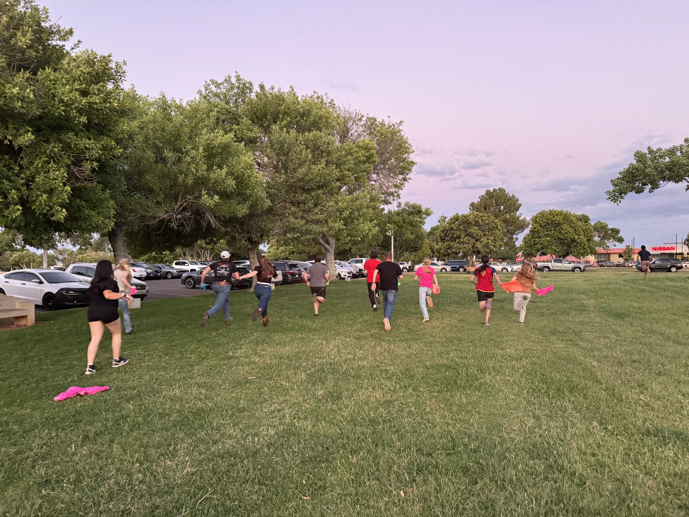 A group of young adults, seen from behind, running across a large grassy field at dusk, with trees and a parking lot full of cars in the background.
