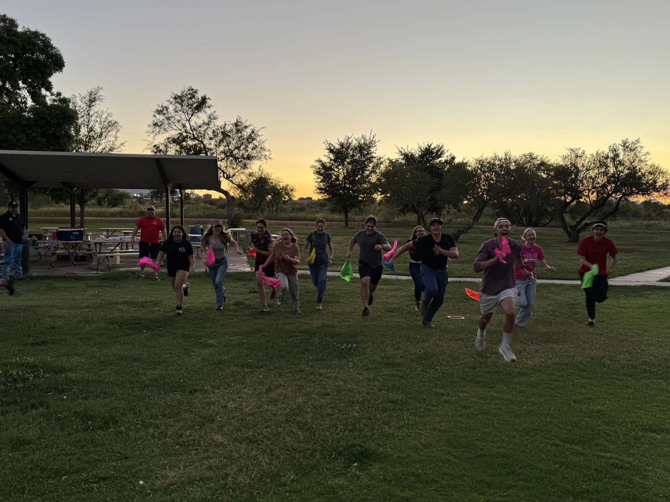 A group of young people run across a grassy field at dusk, smiling and holding colorful flags while playing a game.