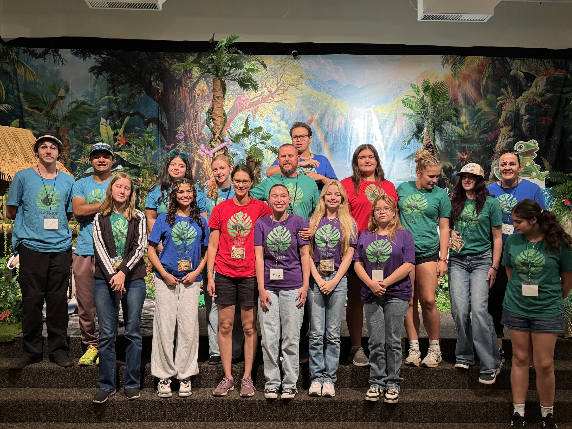 A large group of teenagers and adults in colorful matching shirts stand on steps for a photo in front of a vibrant jungle and waterfall backdrop.