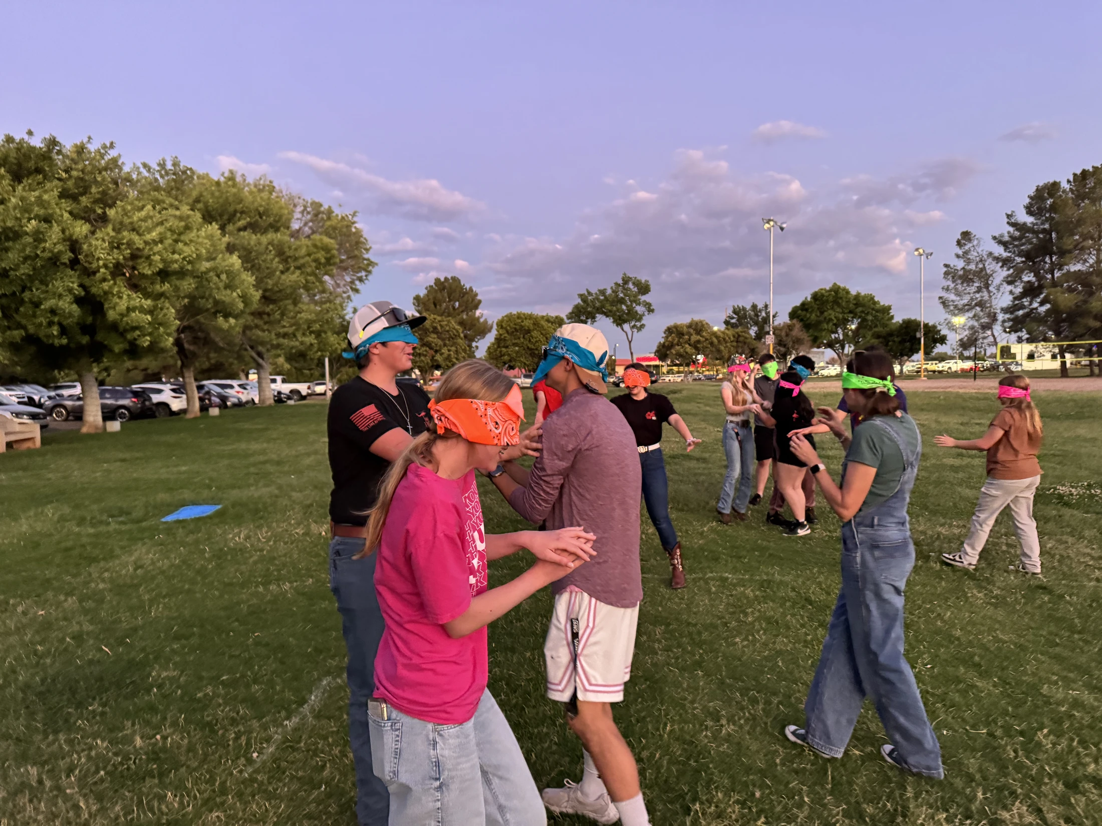 A group of young adults wearing colorful bandana blindfolds play a game on a grassy field at dusk, with arms outstretched as they try to navigate.