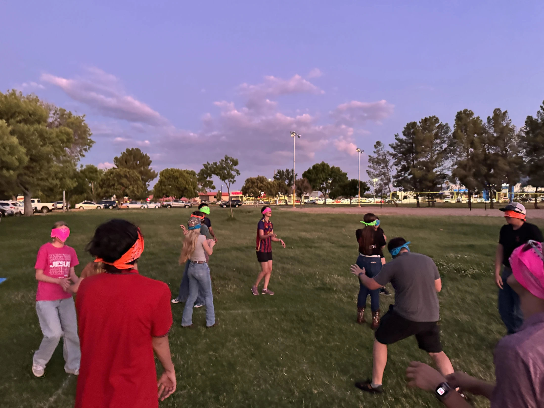 A group of young people wear colorful blindfolds while playing a game on a grassy field at dusk under a purple sky.