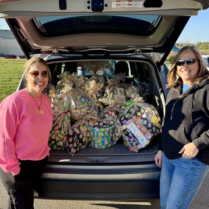 Two smiling women stand in front of the open trunk of an SUV, which is packed full of colorful, cellophane-wrapped Easter baskets for a charity event.
