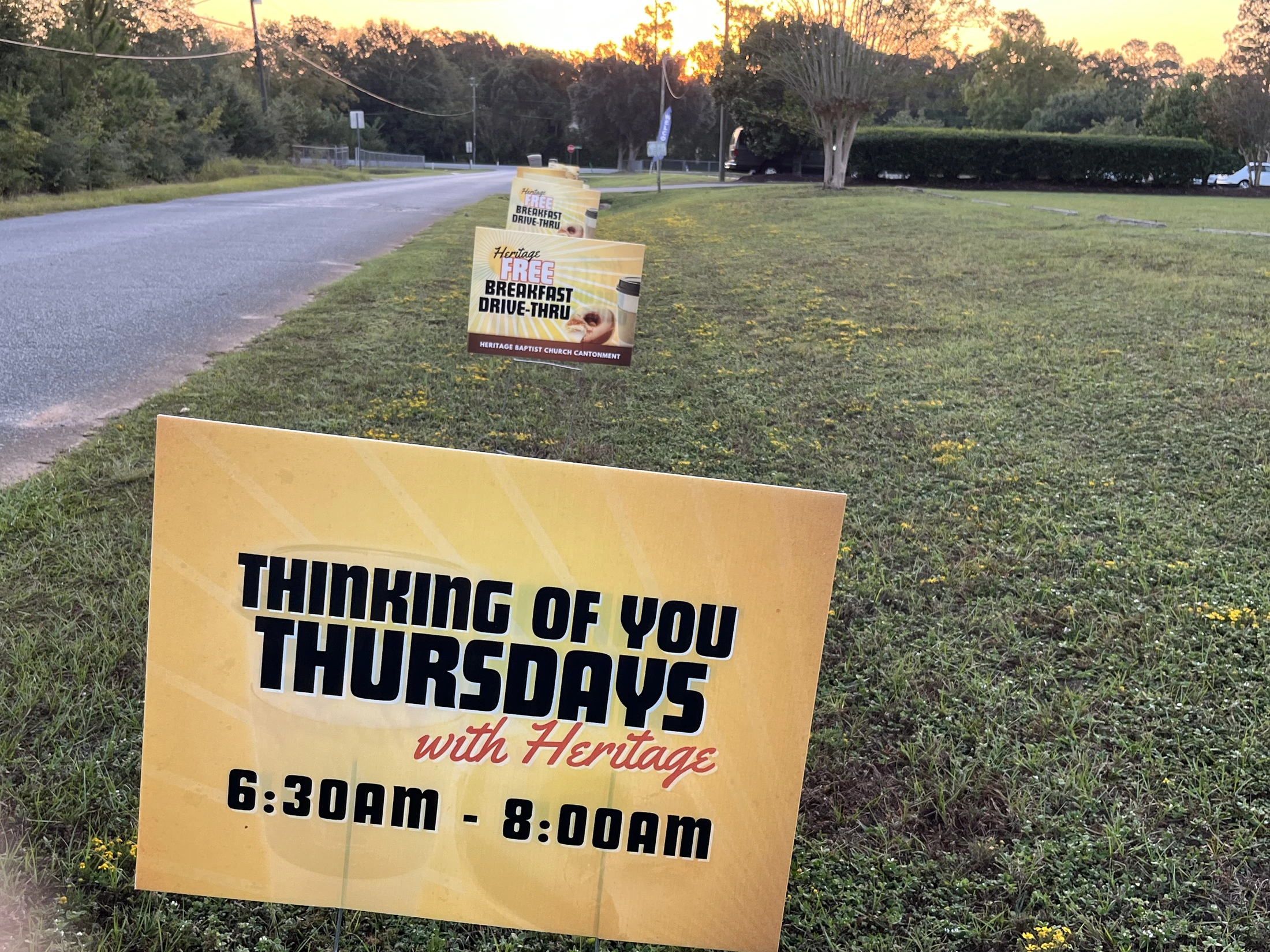 A row of yellow signs on a grassy roadside advertises a free breakfast drive-thru event called "Thinking of You Thursdays" from 6:30 AM to 8:00 AM.