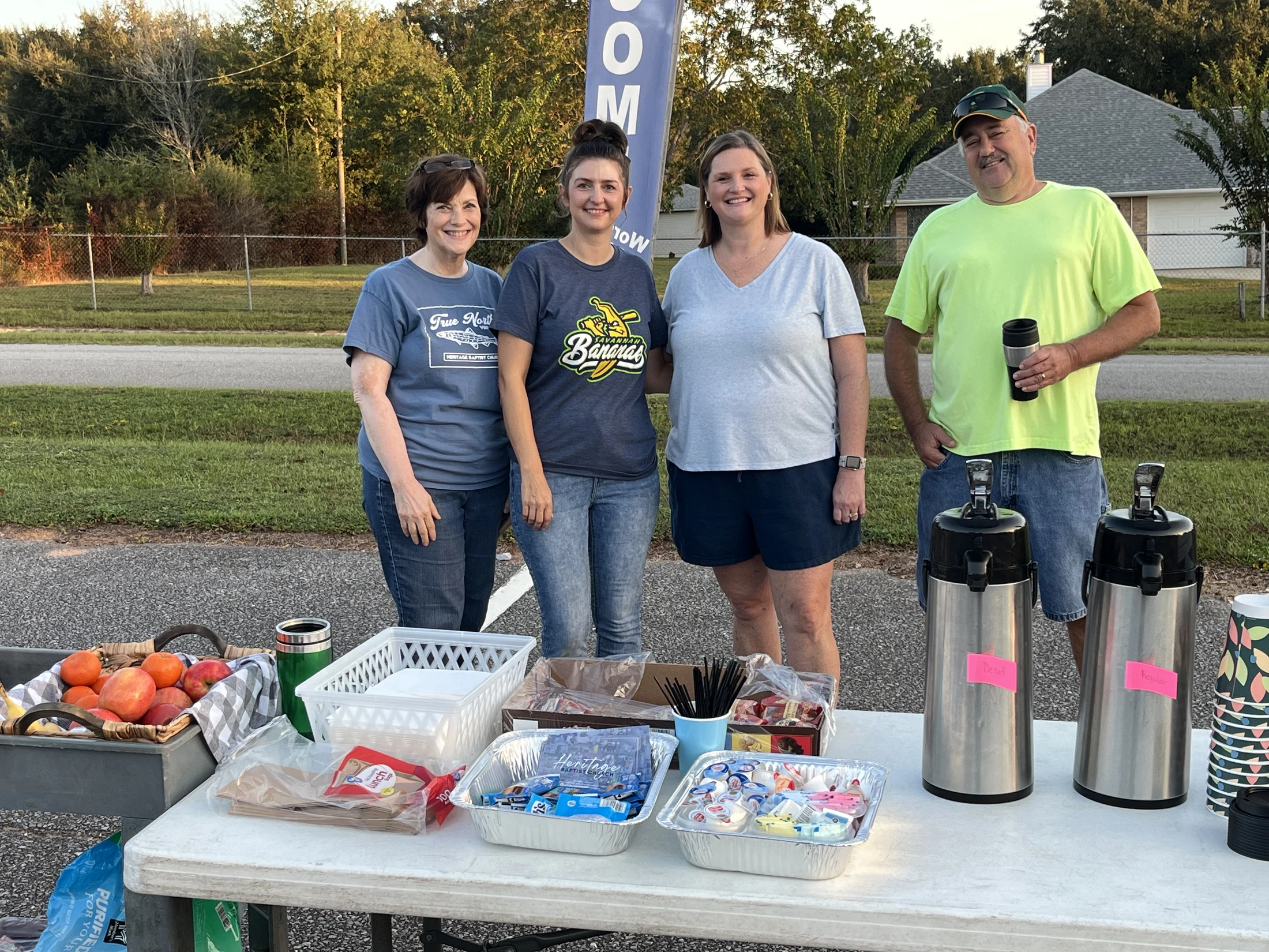 Four smiling volunteers, three women and one man, stand behind a table set up with coffee dispensers, fruit, and various snacks at an outdoor event.