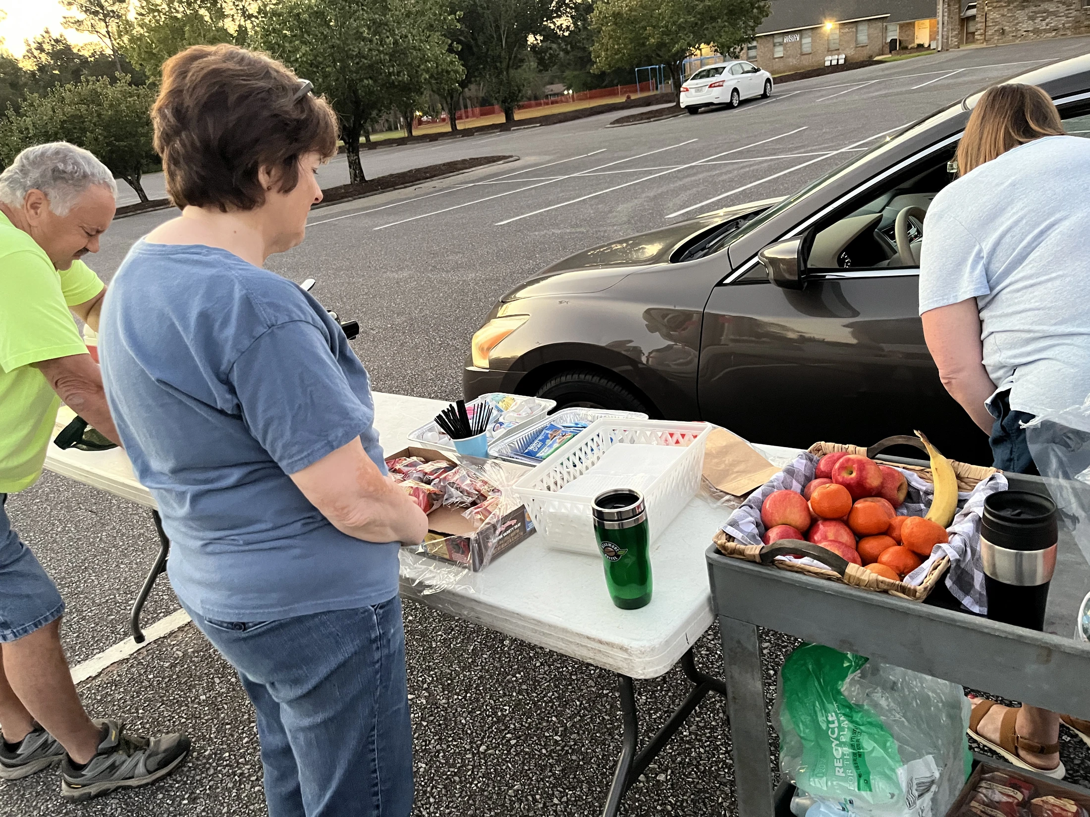 Three people stand by a folding table in a parking lot, set with snacks like fruit and pastries, next to a car at dusk.