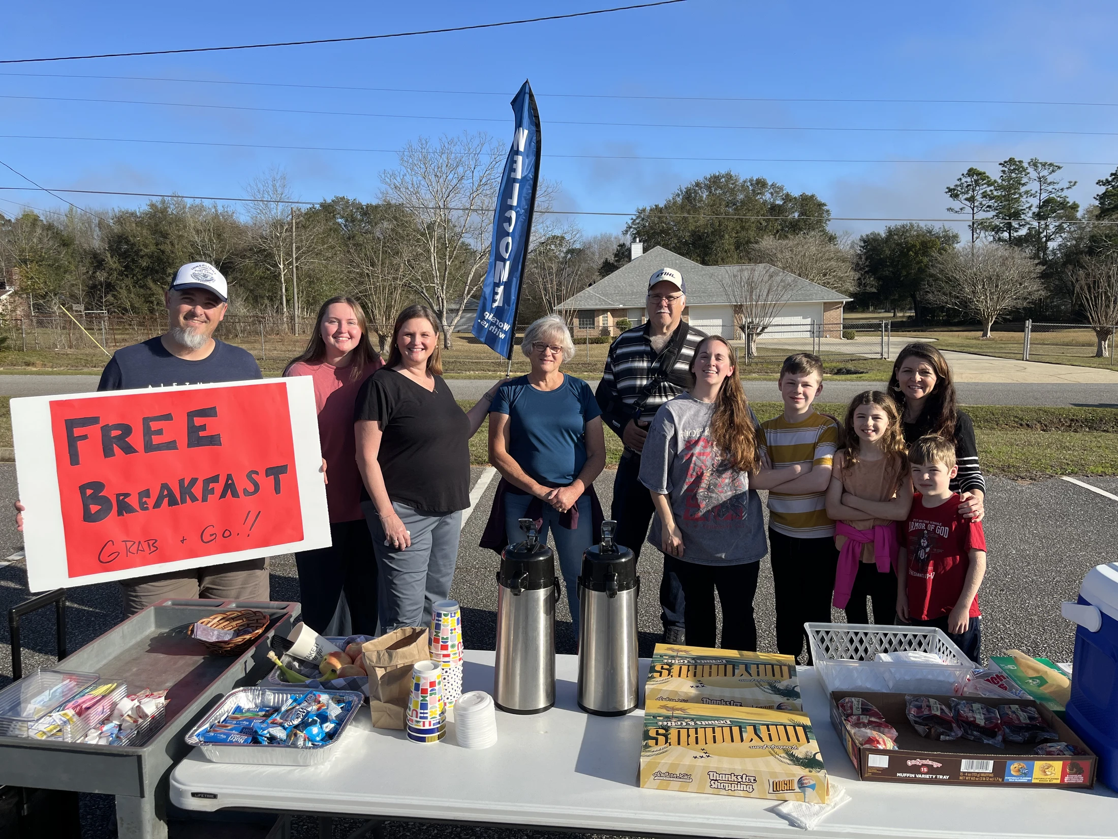 A smiling group of adults and children stand outdoors behind a table with a sign offering a 'Free Breakfast Grab + Go!' to the community.